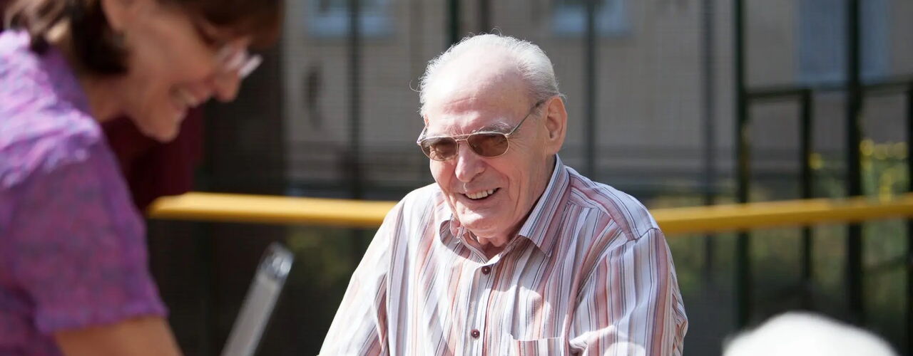Ein älterer, sehbehinderter Mann mit getönter Brille sitzt draußen vor einem gebäude. Er befindet sich im Gespräch mit einer Frau, die ihm gegenübersteht. Beide Personen lächeln.